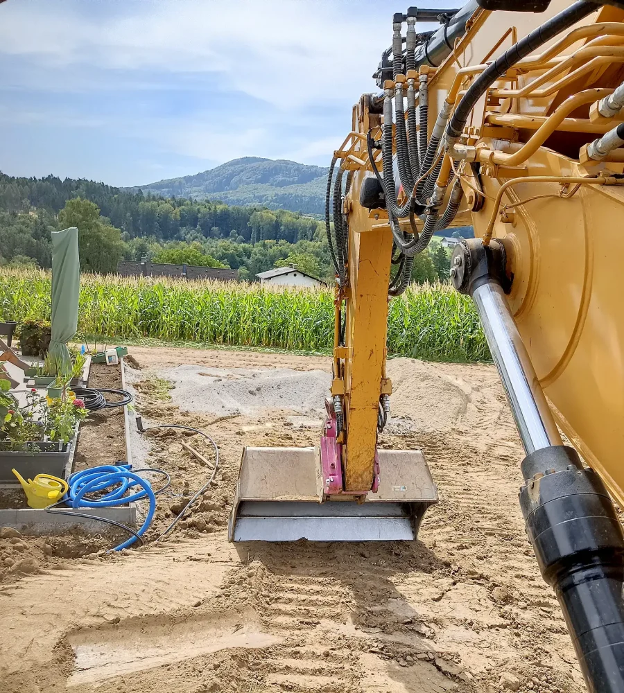 Der Blick aus dem Inneren eines Baufahrzeugs zeigt einen gelben Baggerarm, der sich in Richtung einer sandigen Baustelle ausdehnt. Links ist ein kleiner Garten mit Pflanzen in Töpfen und in der Nähe ist ein blauer Schlauch aufgerollt. Dahinter sind ein Maisfeld und von Bäumen gesäumte Hügel unter einem blauen Himmel zu sehen.